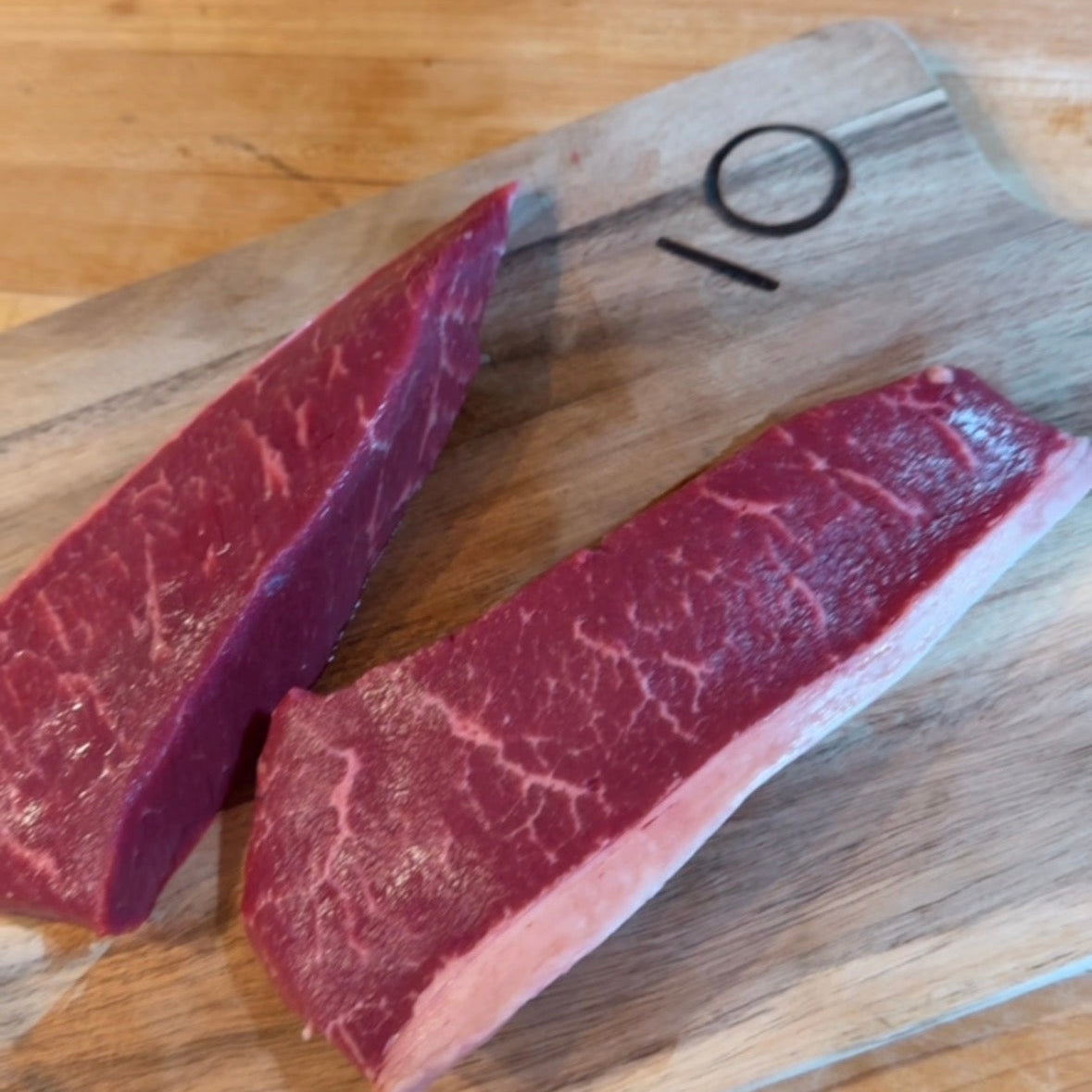 Two pieces of raw beef on a wooden cutting board with a brand logo.