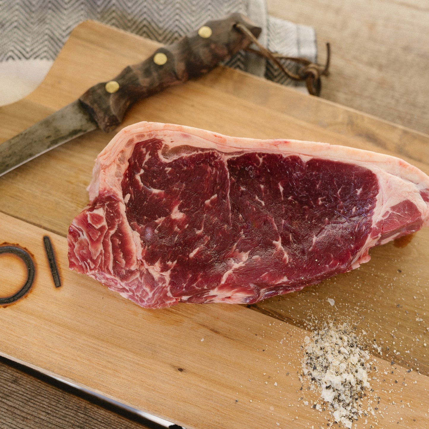 Raw steak on a wooden cutting board with a knife and seasoning.
