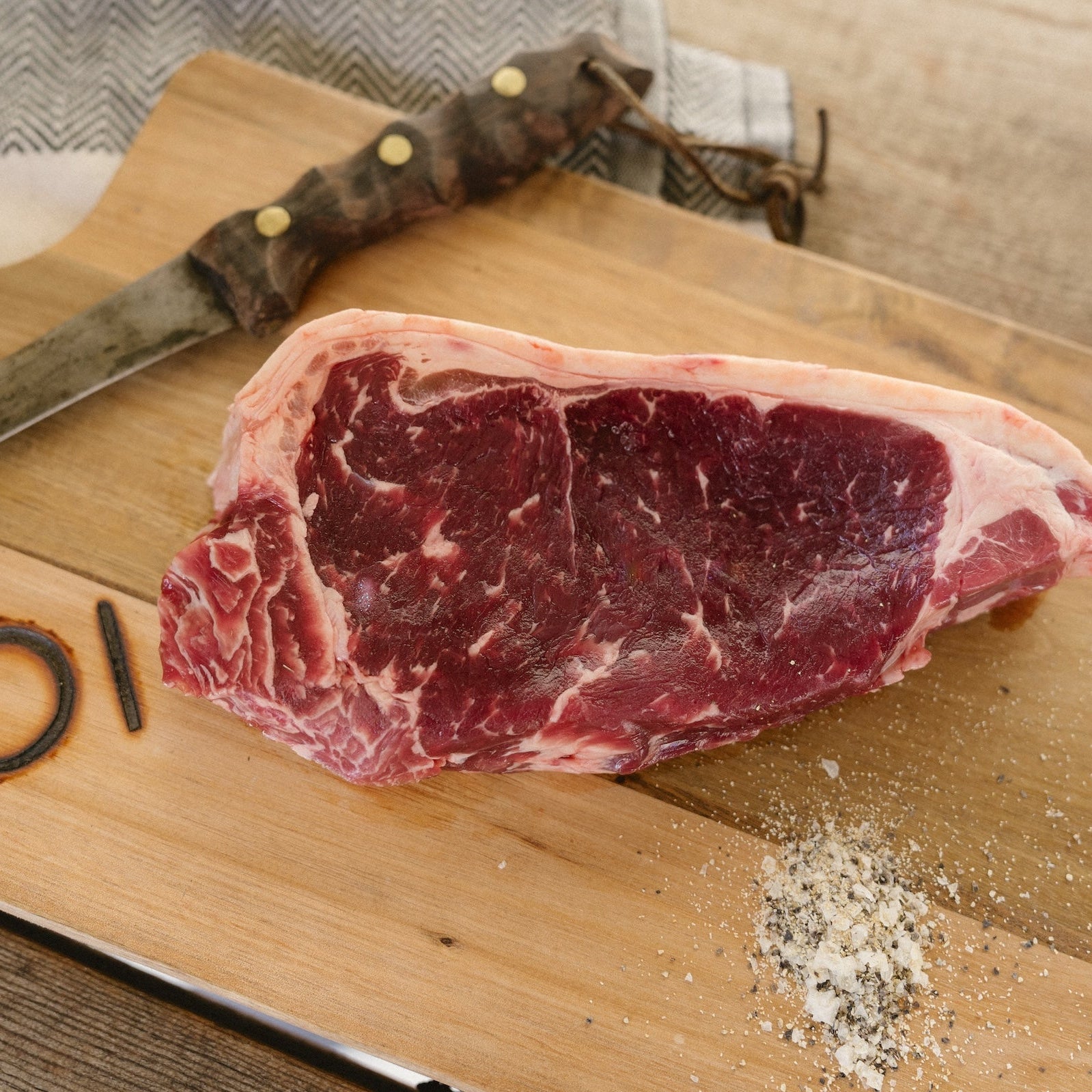 Raw steak on a wooden cutting board with a knife and seasoning.